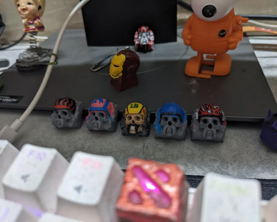 A row of painted skull keycaps in different helmet colours, with more keycaps on the desk behind
