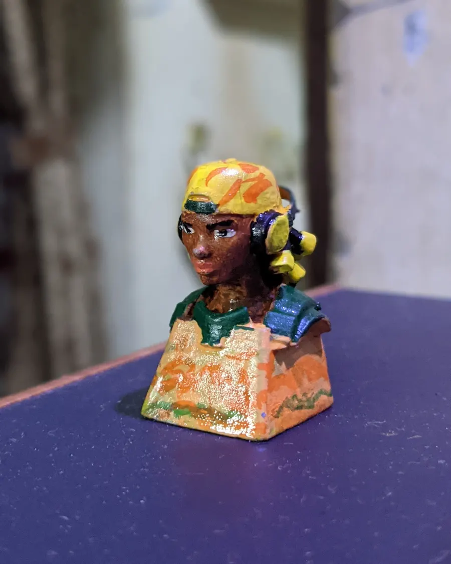 The Raze: a small hand-painted bust of a character in a yellow cap and headphones, sitting on a keycap base